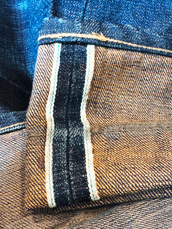 Close-up of a denim garment showing the inner leg cuff with a striped contrast stitching detail in dark navy, beige, and white on a brown-toned twill fabric.