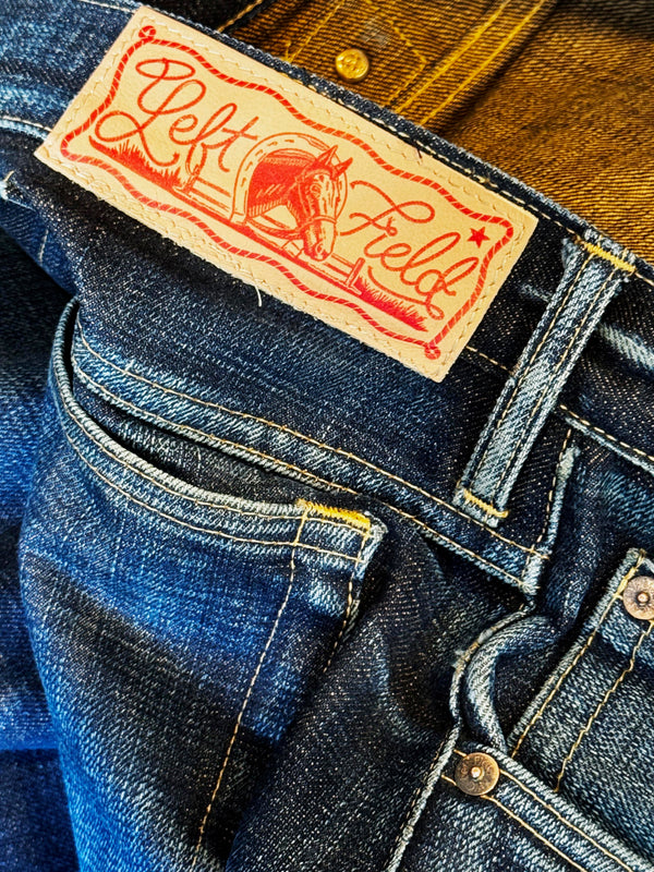 Close-up of dark blue denim jeans showing the waistband, pocket, and a tan leather brand patch with red script reading 'Left Field'.