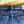 Close-up of the back waistband of dark blue denim jeans with a matching belt, showing a metal buckle, copper-toned rivets, and a pink brand label on a yellow wooden background.