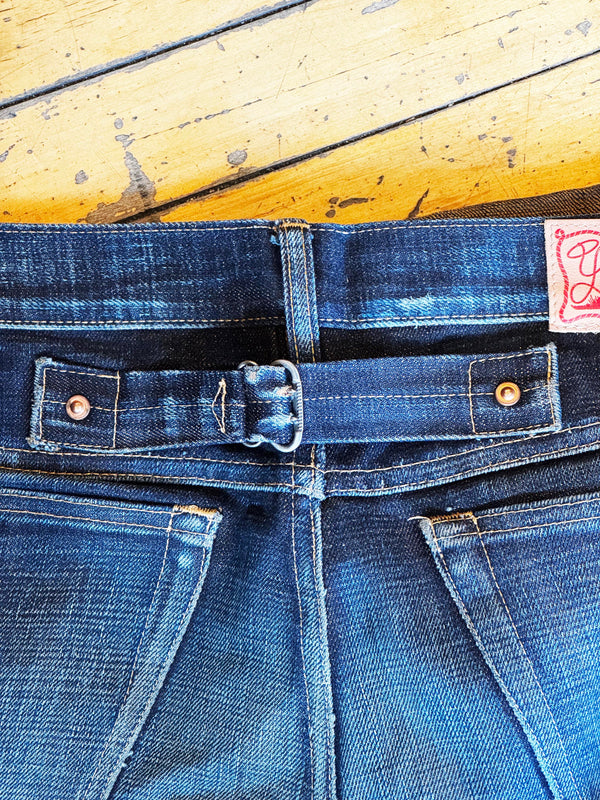 Close-up of the back waistband of dark blue denim jeans with a matching belt, showing a metal buckle, copper-toned rivets, and a pink brand label on a yellow wooden background.