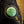 A close-up of a circular vintage-style metal button or badge with a green patina and a hammer-and-sickle symbol, set against dark textured fabric with gold-toned stitching.
