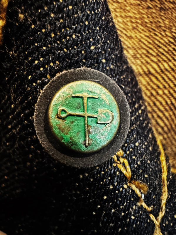 A close-up of a circular vintage-style metal button or badge with a green patina and a hammer-and-sickle symbol, set against dark textured fabric with gold-toned stitching.