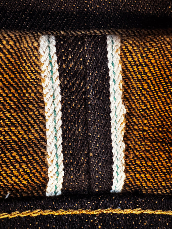 Close-up of a textured denim fabric showing a classic diagonal twill weave in dark brown and golden yellow, accented by two vertical white stripes with subtle green and blue threads.