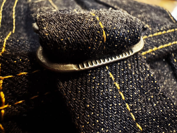 Close-up of a rolled dark indigo denim fabric with bright yellow contrast stitching and a small metal clip or fastener attached near the top center.