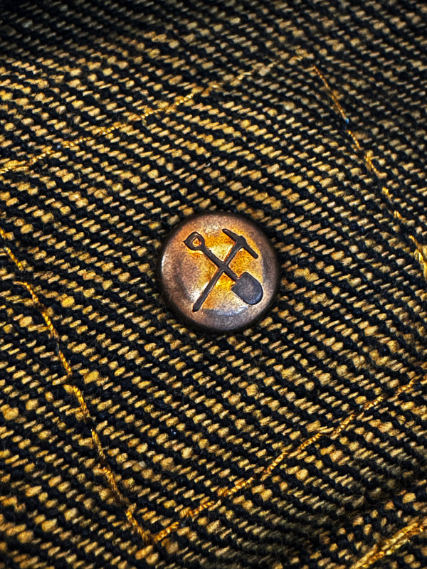 A close-up of a round metal button with a distressed bronze finish, featuring an engraved crossed pickaxe and shovel symbol, set against a textured black and gold woven denim background.