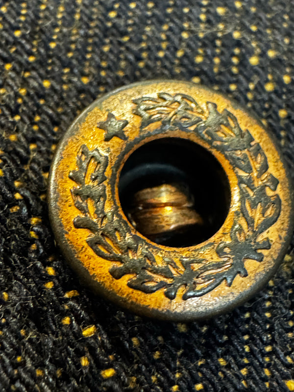 A close-up of a round brass button with an engraved laurel wreath and star pattern, set against a black textured fabric background with yellow stitching.