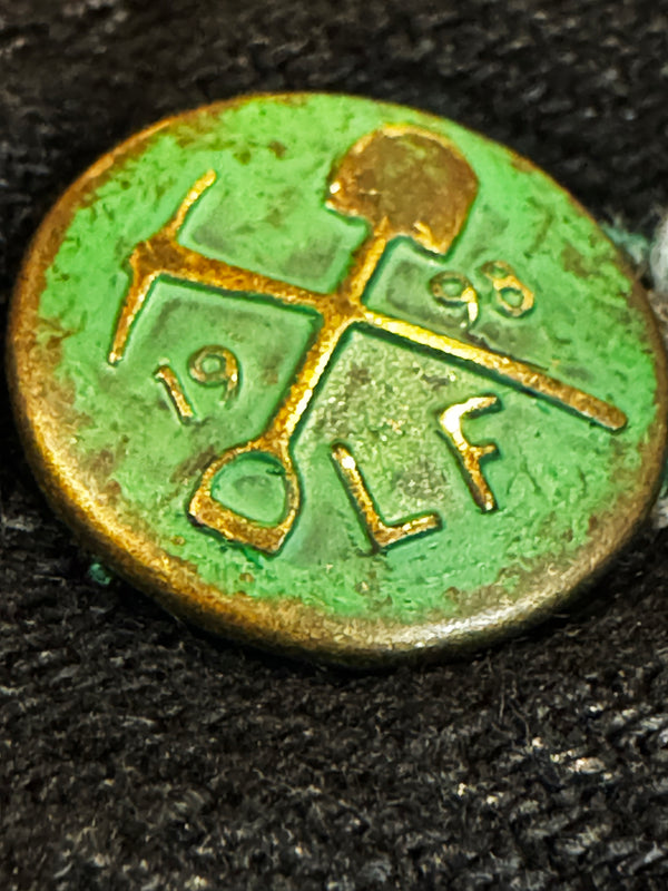 A close-up of a round, antique-style metal button or medallion with a green patina and gold-toned lettering and symbols, resting on a dark textured fabric background.