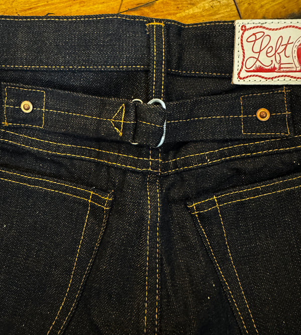Close-up of the back waistband of dark indigo denim jeans, showing a belt loop, button, yellow contrast stitching, and a white brand patch with red script reading 'Left'.