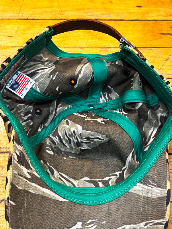 Close-up of the inside of a camouflage baseball cap with bright teal trim and a brown leather strap, laid on a yellow wooden surface.