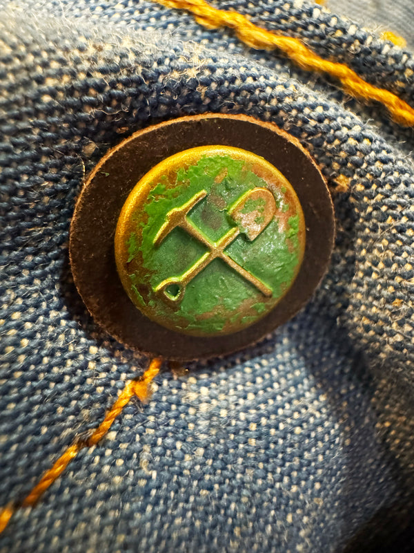 A close-up of a round vintage-style metal button with crossed tools symbol, mounted on blue denim fabric with orange stitching.