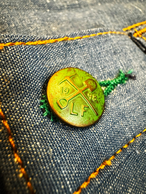 A close-up of a round vintage brass button with '1946 OLF' engraved, pinned onto a blue denim pocket with orange contrast stitching.