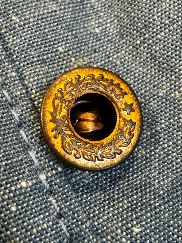A close-up of a round antique brass button with engraved floral details, shown on a textured blue denim background.
