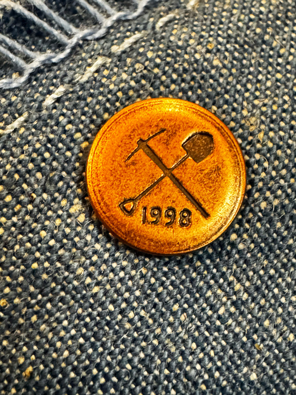 A round copper-colored metal button with crossed tools and the year 1998, shown on a textured gray denim fabric background.