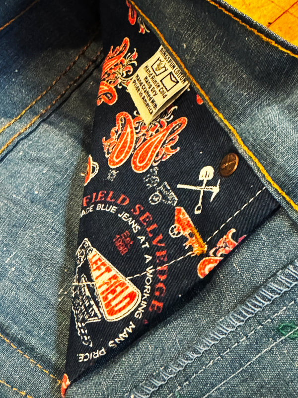 Close-up of the inner neck lining of a pair of blue jeans, showing the 'Field Selvage' brand label with red and white graphics on a black background, alongside the button and denim fabric.