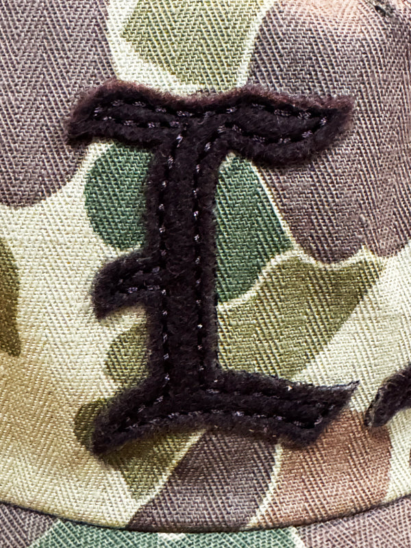 Close-up of a camouflage fabric patch with a large dark brown embroidered letter 'I'.