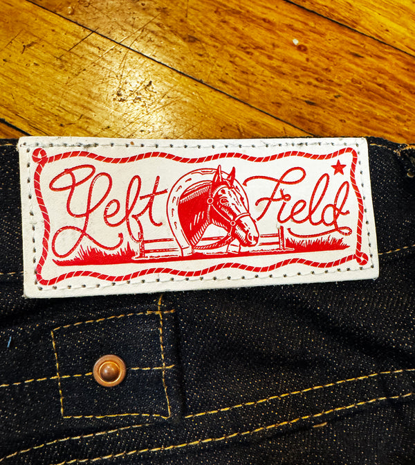 A close-up of a denim garment showing a white rectangular brand patch with red embroidery that reads 'Left Field' and includes a horse illustration. The patch is sewn onto dark indigo denim, with a wooden floor visible in the background.